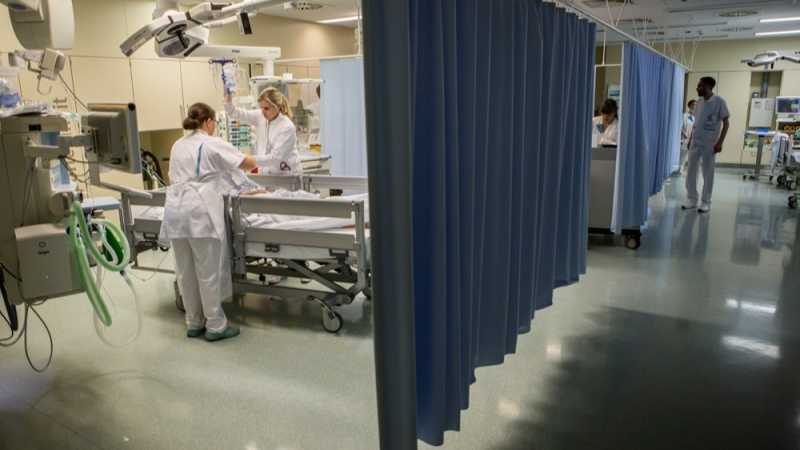 Das Bild zeigt eine Krankenhausstation mit mehreren, durch einen Vorhang abgetrennten, Betten. Der Blick richtet sich auf ein Krankenbett links im Bild, an dem eine Ärztin und eine Pflegerin einen Patienten versorgen.