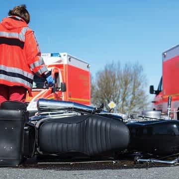 Man sieht ein beschädigtes Motorrad auf dem Boden liegen, im Hintergrund läuft eine Sanitäterin zu einem Rettungswagen.