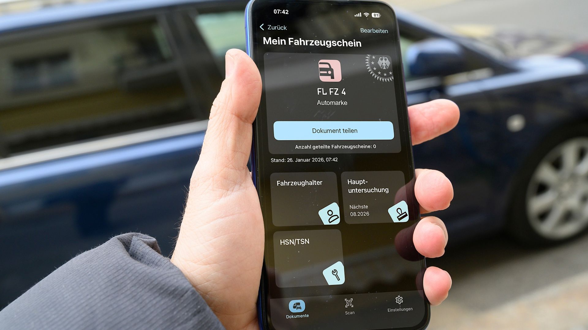 Eine Hand hält ein Handy, auf dem die App „Mein Fahrzeugschein“ geöffnet ist. Die App zeigt Fahrzeugdaten, Fahrzeughalter und Hauptuntersuchung an.