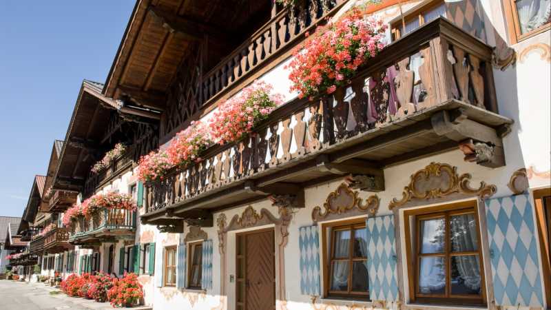 das Bild zeigt ein bayrisches Fachwerkhaus mit Holzbalkon, Blumenkästen und blau-weiß karierten Fensterläden.