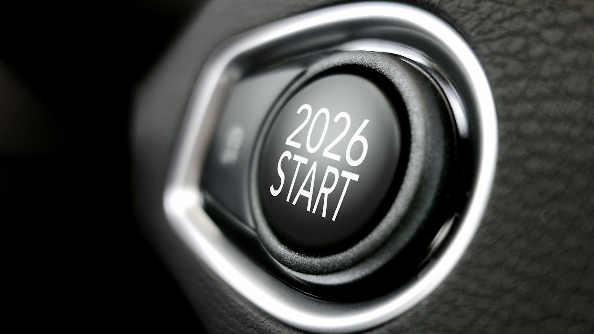 Ein schwarzer Startknopf in einem Kraftfahrzeug, auf dem „2026 Start“ steht. Der Knopf ist von einem glänzenden, silberfarbenen Ring eingefasst und vor einem dunklen, genarbten Hintergrund platziert.