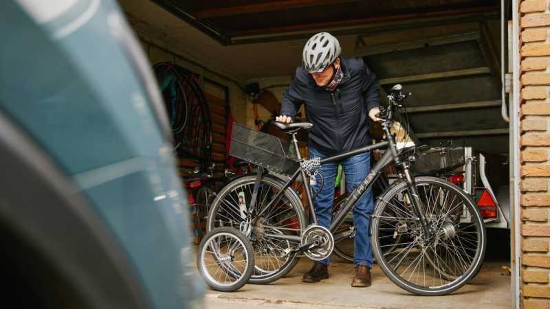 Ein Mann mit Fahrradhelm steht in einer Garage und beugt sich über ein schwarzes Fahrrad mit Stützrädern.