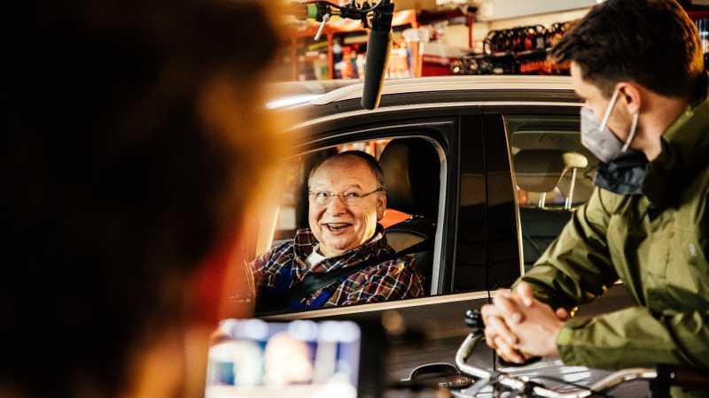 Ein Foto vom Set des Drehs mit Gernot Hassknecht. Hassknecht sitzt im Auto und lächelt einem Mann zu, der neben ihm auf einem Fahrrad sitzt. Es sind Kameras, Mikrofone und weitere Film-Utensilien zu sehen. 