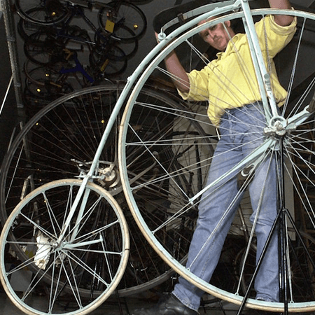 Ein Mann präsentiert ein historisches Hochrad mit übergroßem Vorderrad und weißem Rahmen.