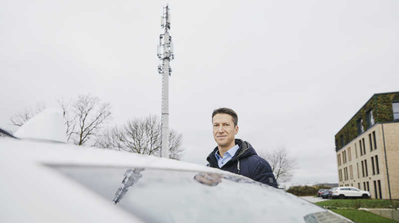 Portrait von Prof. Christoph Degen. Er steht neben seinem Auto auf einem Parkplatz, hinter ihm ist ein Mobilfunkmast zu sehen.