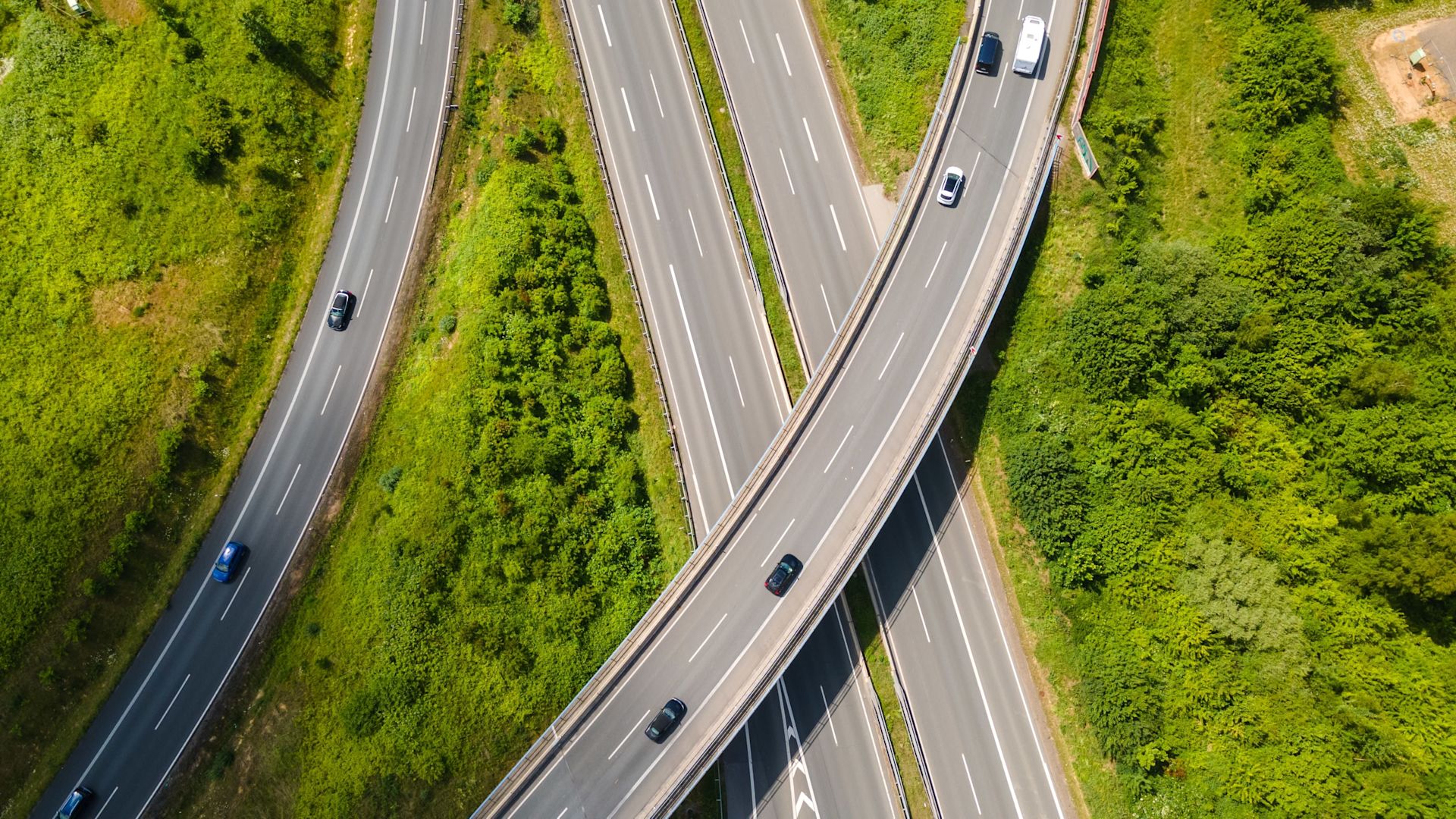 Luftaufnahme einer Autobahngabelung mit Brücken, Straßen und fahrenden Autos, umgeben von grünen Bäumen und Wiesen.