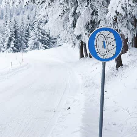 ein Straßenschild für Schneekettenpflicht steht neben einer stark eingeschneiten Straße.