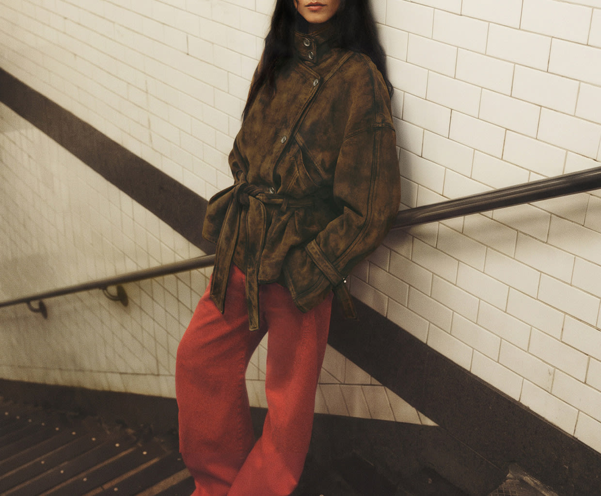 Woman with long dark hair wearing brown suede jacket and red pants leaning against white tiled subway wall with handrail.