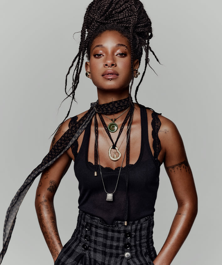 Woman with long braided hair wearing black tank top and layered pendant necklaces against light background