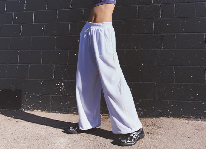 Person wearing light purple wide-leg sweatpants and black sneakers standing against a dark brick wall in sunlight.