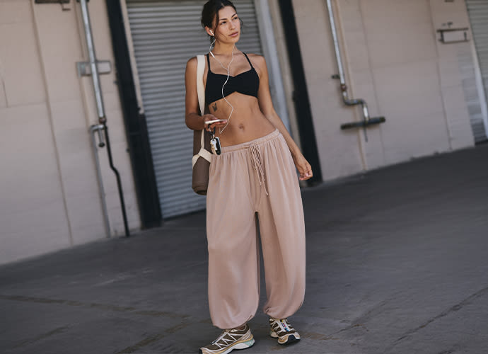 Woman in black crop top and beige wide-leg pants holding sunglasses, standing outdoors near industrial building.