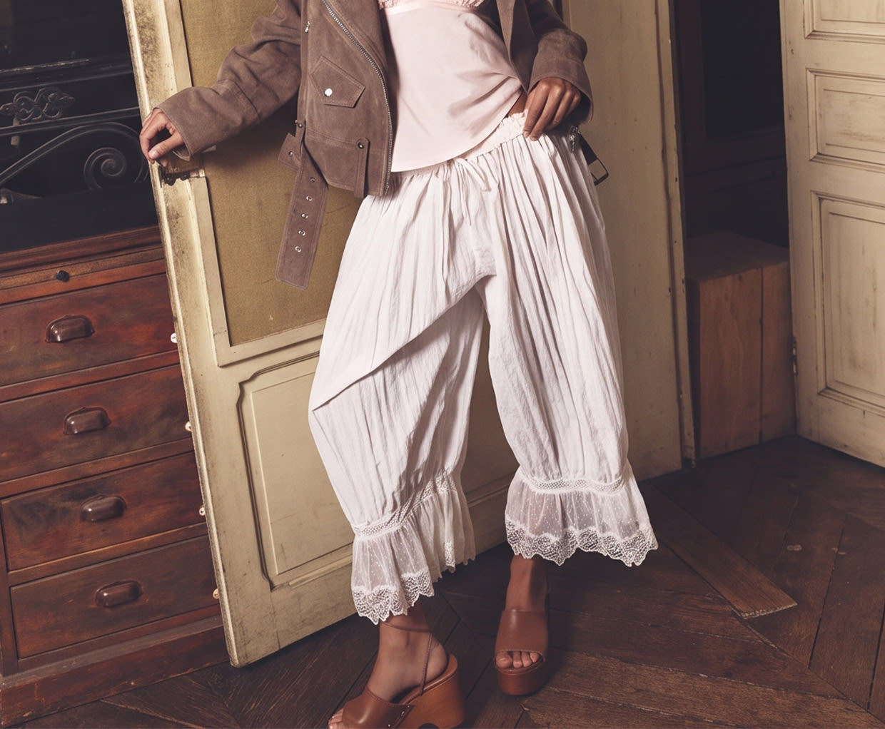 Woman in brown jacket and white lace-trimmed bloomers standing by vintage wooden dresser in rustic interior.