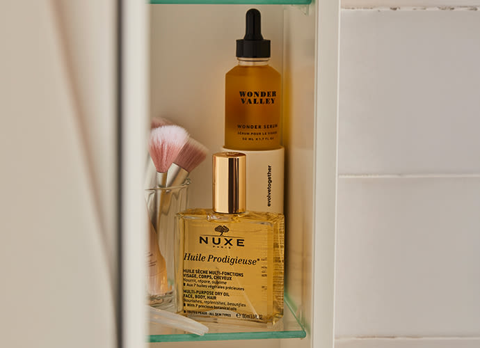 Glass shelf displaying beauty products including NUXE Huile Prodigieuse oil, Wonder Valley serum, and pink makeup brushes.