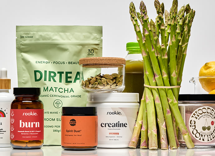 Assorted health and wellness products including matcha tea, supplements, and fresh asparagus arranged on a white surface.