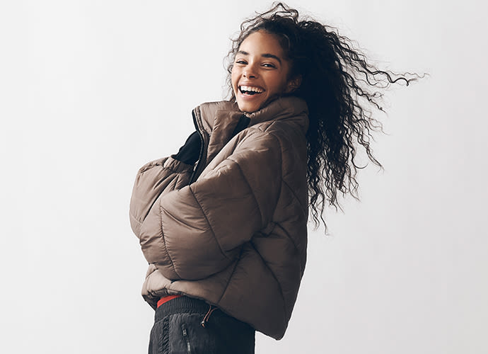 Young woman with long curly black hair laughing joyfully, wearing a brown puffer jacket against a white background.