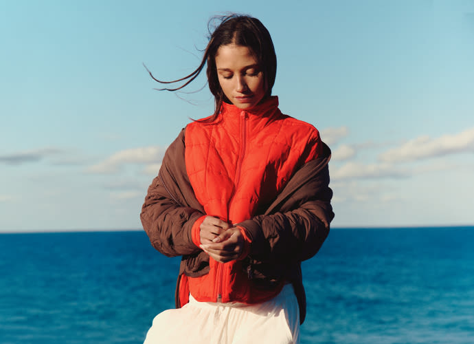 Woman in red and brown jacket standing by blue ocean with hands clasped, looking down peacefully under clear sky.