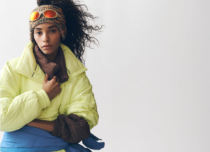 Woman wearing yellow puffer jacket and knit beanie with goggles, sitting against white background with windswept hair.