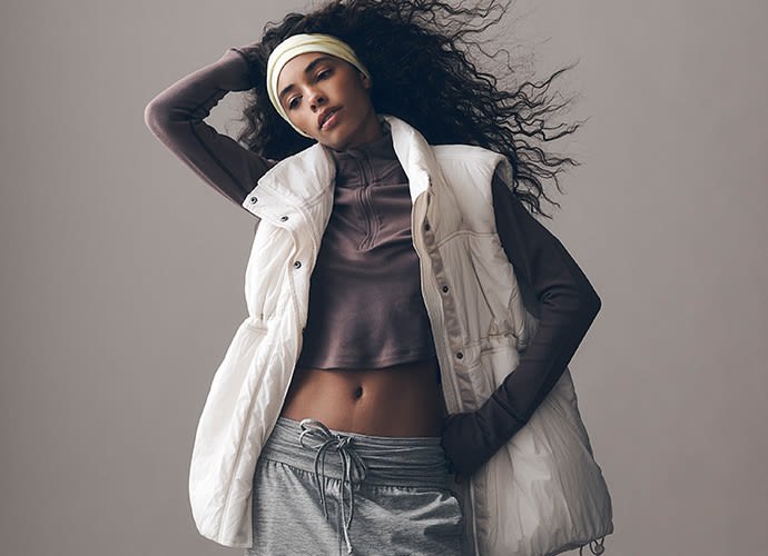 Woman in white vest and headband with flowing curly hair, wearing athletic wear against neutral background.