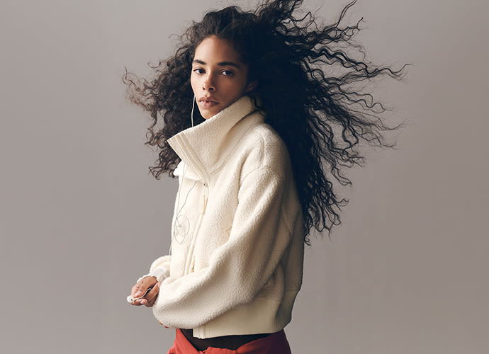 Woman with long curly black hair flowing in motion, wearing a cream-colored fleece jacket against a neutral gray background.