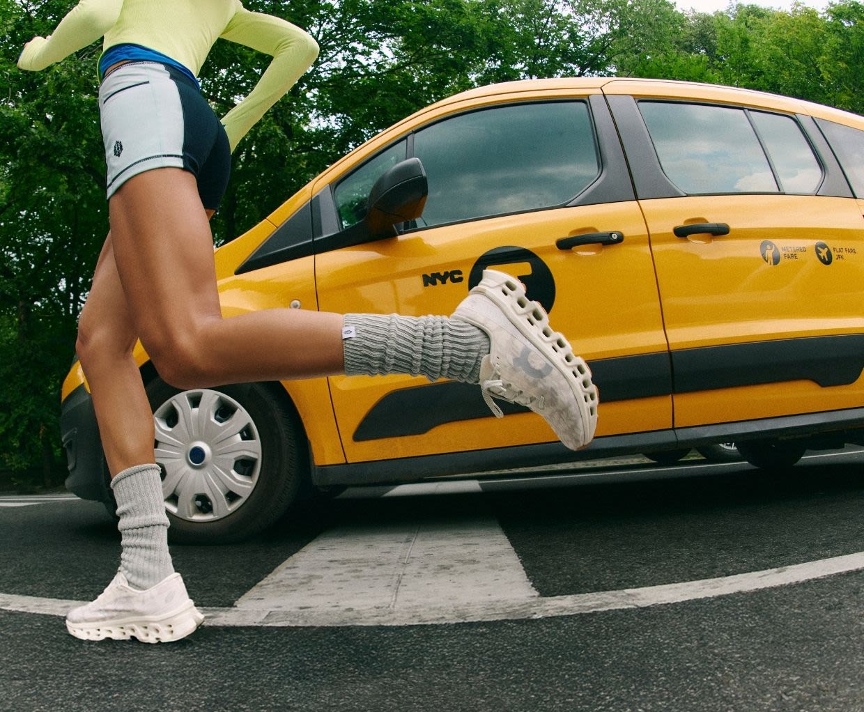 Runner in white sneakers and athletic wear jogging past a yellow NYC taxi cab with green trees in the background.