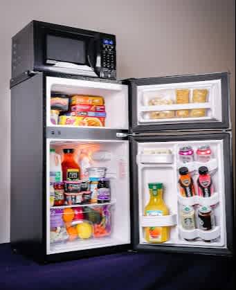 A microwave on top of a mini-fridge with freezer and refrigerator sections filled with food and beverages.
