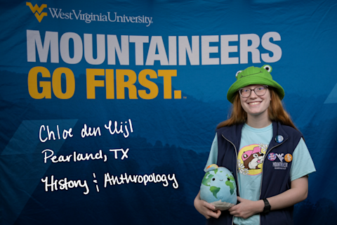 Chloe den Uijl, a History and Anthropology major from Pearland, Texas, wears a green frog hat and blue Mountaineer Marching Band vest, and holds a stuffed globe toy.