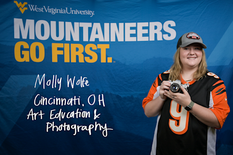 Molly Wolfe holds a camera while wearing an orange and black Bengals jersey and hat. She is an Art Education and Photography major from Cincinnati, Ohio.