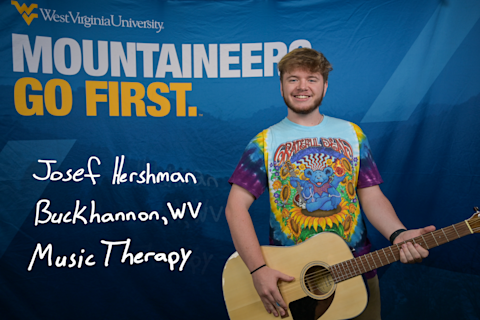 Josef Hershman stands in front of the blue Mountaineers Go First backdrop holding a guitar. He is wearing a Grateful Dead T-shirt. Josef is a Music Therapy major from Buckhannon, WV.