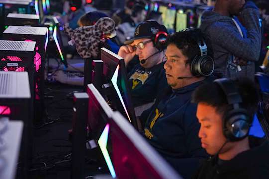 WVU esports team members compete in a darkened room. They are wearing headsets and are seated in front of computer monitors.