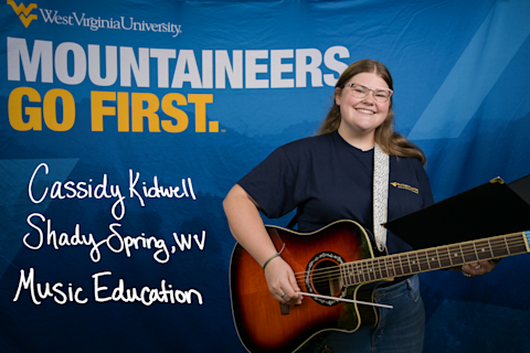 Cassidy Kidwell, a Music Education major from Shady Spring, WV, wears a guitar on a shoulder strap and holds a conducting baton and an open folder.