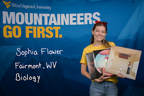 Sophia Flower, a Biology major from Fairmont, WV, holds three albums. She is wearing a gold shirt and jeans, and has a pair of heart-shaped red sunglasses on her head.