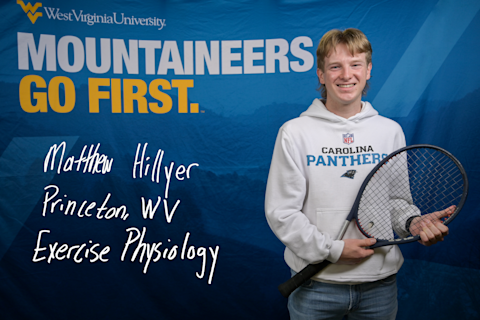 Matthew Hillyer stands in front of the blue Mountaineers Go First backdrop. He wears a white Carolina Panthers sweatshirt and holds a tennis racket. He is an Exercise Physiology major from Princeton, WV.