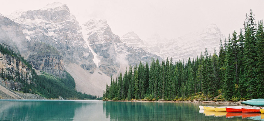 A photo of a lake in front of beautiful mountains. The water is bright blued and there are canoes lined up in the water.