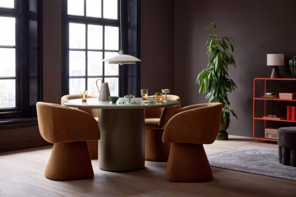 Ronde olijfgroene tafel met oranje stoelen in een stijlvolle kamer, plant, oranje kast en grote ramen op de achtergrond.