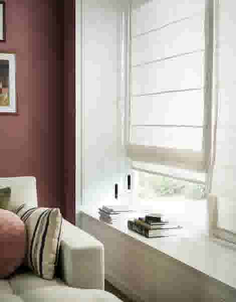 Witte vouwgordijnen hangend voor een raam in een woonkamer met lichte bank en donkerrode muren