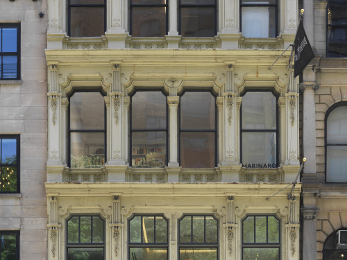The exterior of Marinaro Gallery as seen from ground level on Broadway. The building is an aged yellow concrete with tall skinny windows four across, ornamented with molding and and intricate ribbon details.