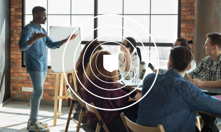 man stood next to a whiteboard leading a group of people sat around a table