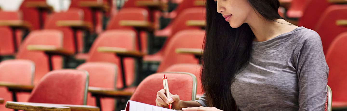 A female test taker in maroon t-shirt sits  in a classroom lecture taking notes for IELTS test preparation.
