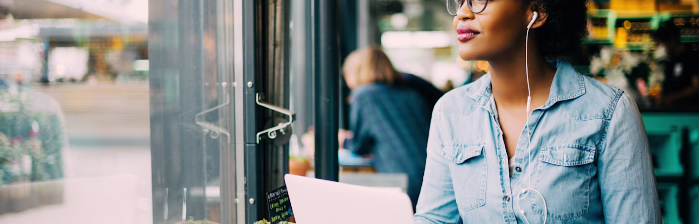 A woman wearing a denim shirt sits near a window and practice for IELTS Listening test on her laptop