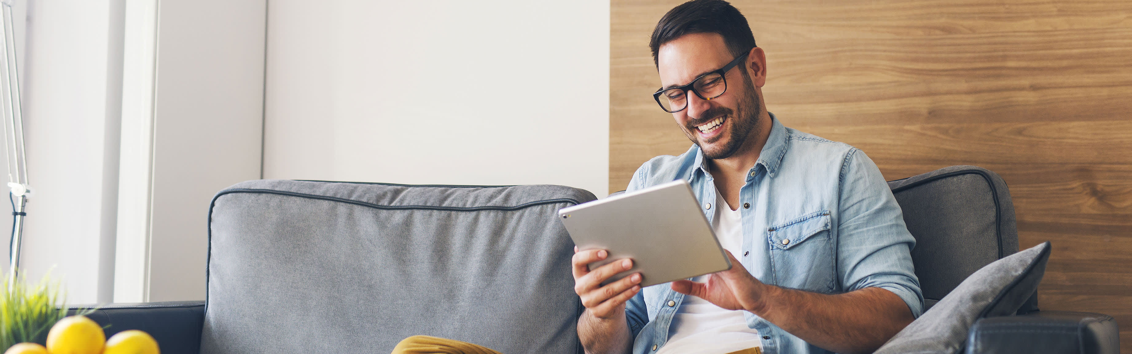 Junger Mann mit Brille sitzt auf Sofa und schaut sich das EnBW DSL-Angebot am Tablet an