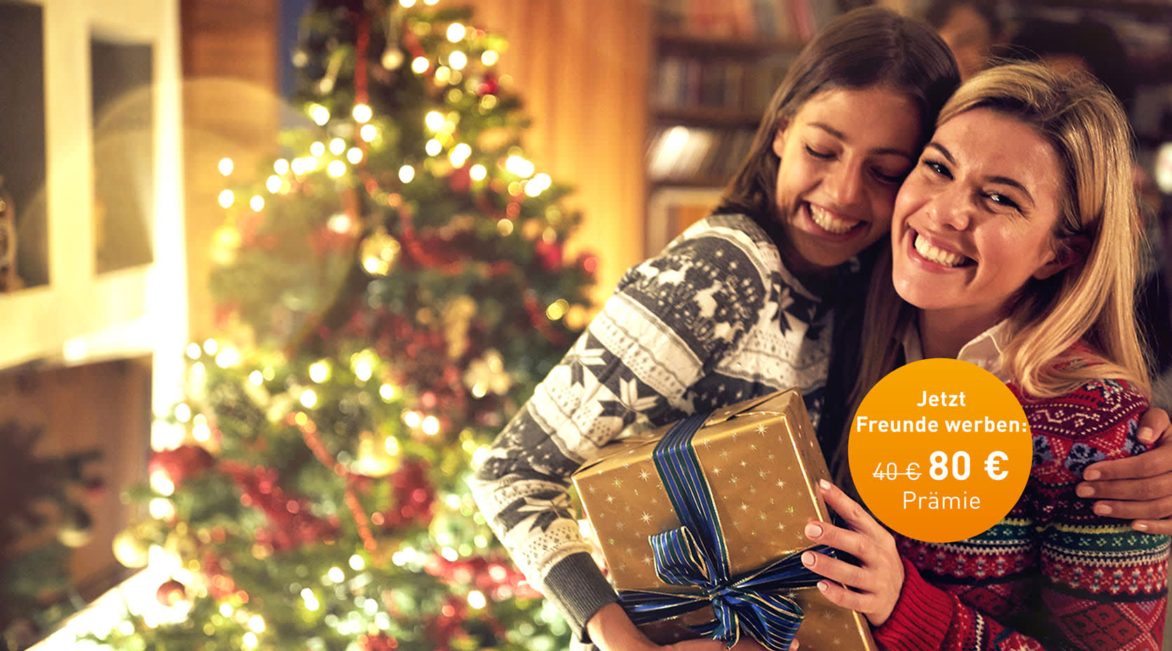 Freundinnen mit Geschenk umarmen sich vor einem Weihnachtsbaum