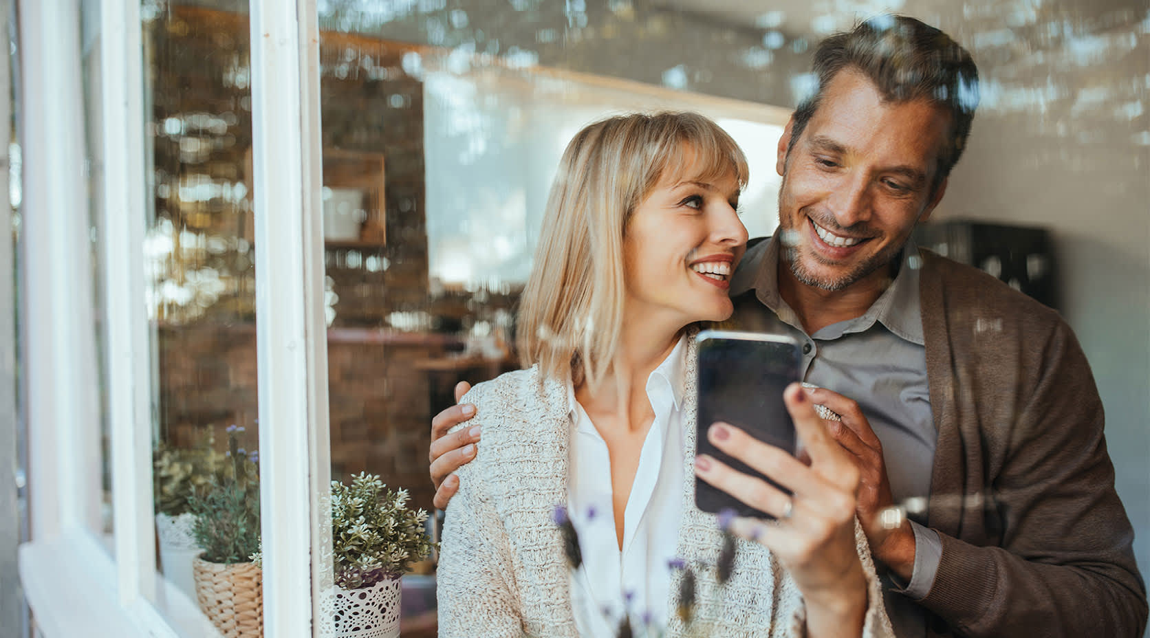 Mann und Frau stehen am Fenster und schauen aufs Handy