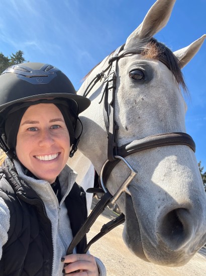Tight shot of woman wearing helmet alongside a gray horse in an English bridle