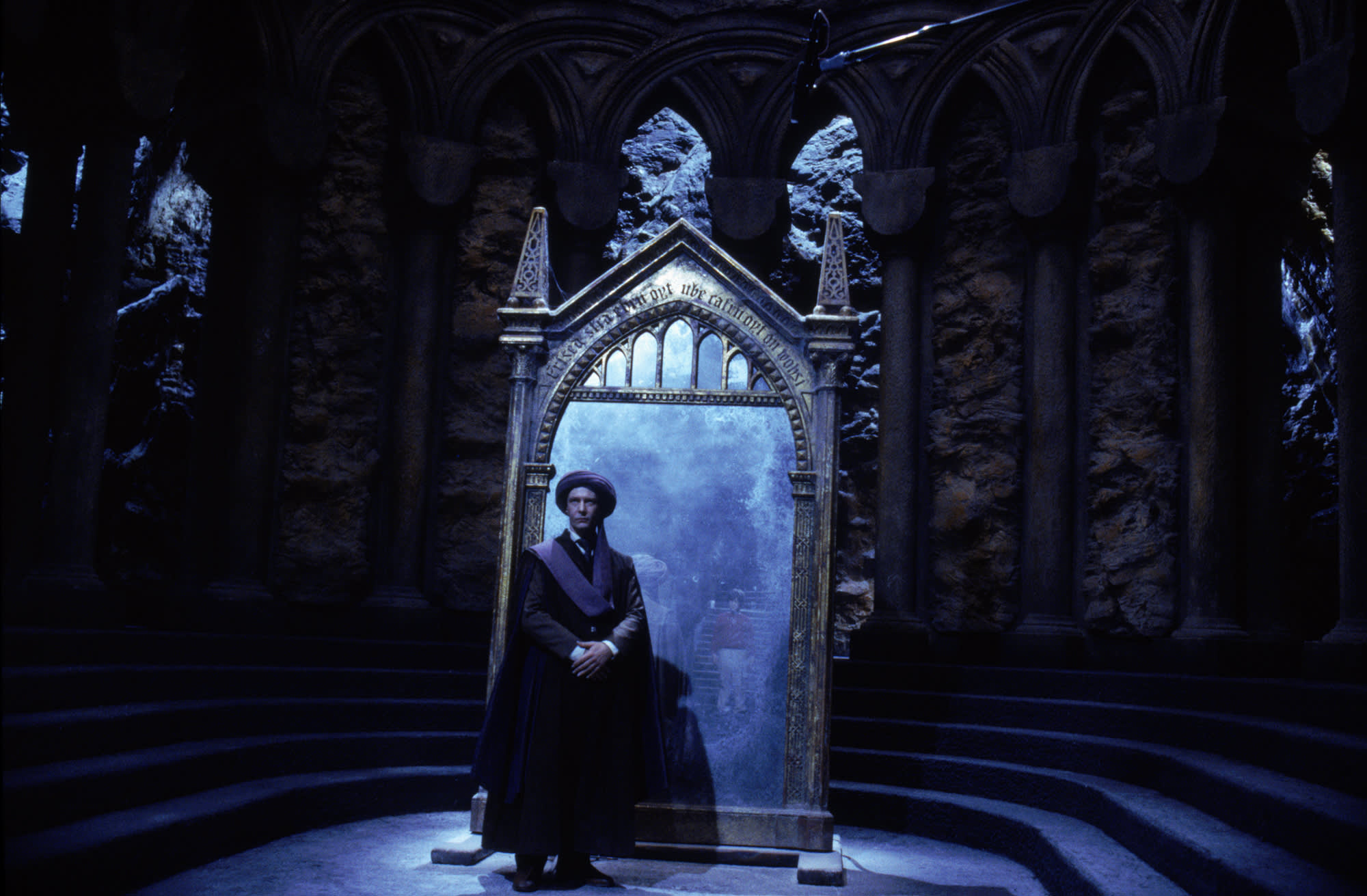 Professor Quirrell is standing with his back to the Mirror of Erised in a stone room.