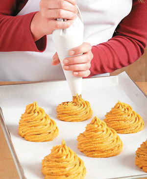 Piping mashed sweet potatoes onto a sheet pan lined with parchment paper using a piping bag.