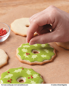 Pipe and fill wreath cookies. While icing is still wet, pipe small ovals onto wreath with lighter green icing. Immediately drag a toothpick to points from ovals, making leaf shapes. Clean toothpick after each drag. Sprinkle wreath with red candy balls.