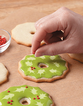 Pipe and fill wreath cookies. While icing is still wet, pipe small ovals onto wreath with lighter green icing. Immediately drag a toothpick to points from ovals, making leaf shapes. Clean toothpick after each drag. Sprinkle wreath with red candy balls.