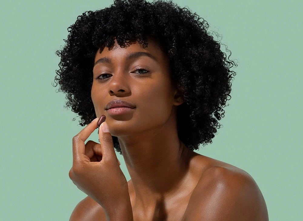 Une femme aux cheveux afro tenant une capsule RICHELET Cheveux, Peau, Ongles entre son pouce et son index