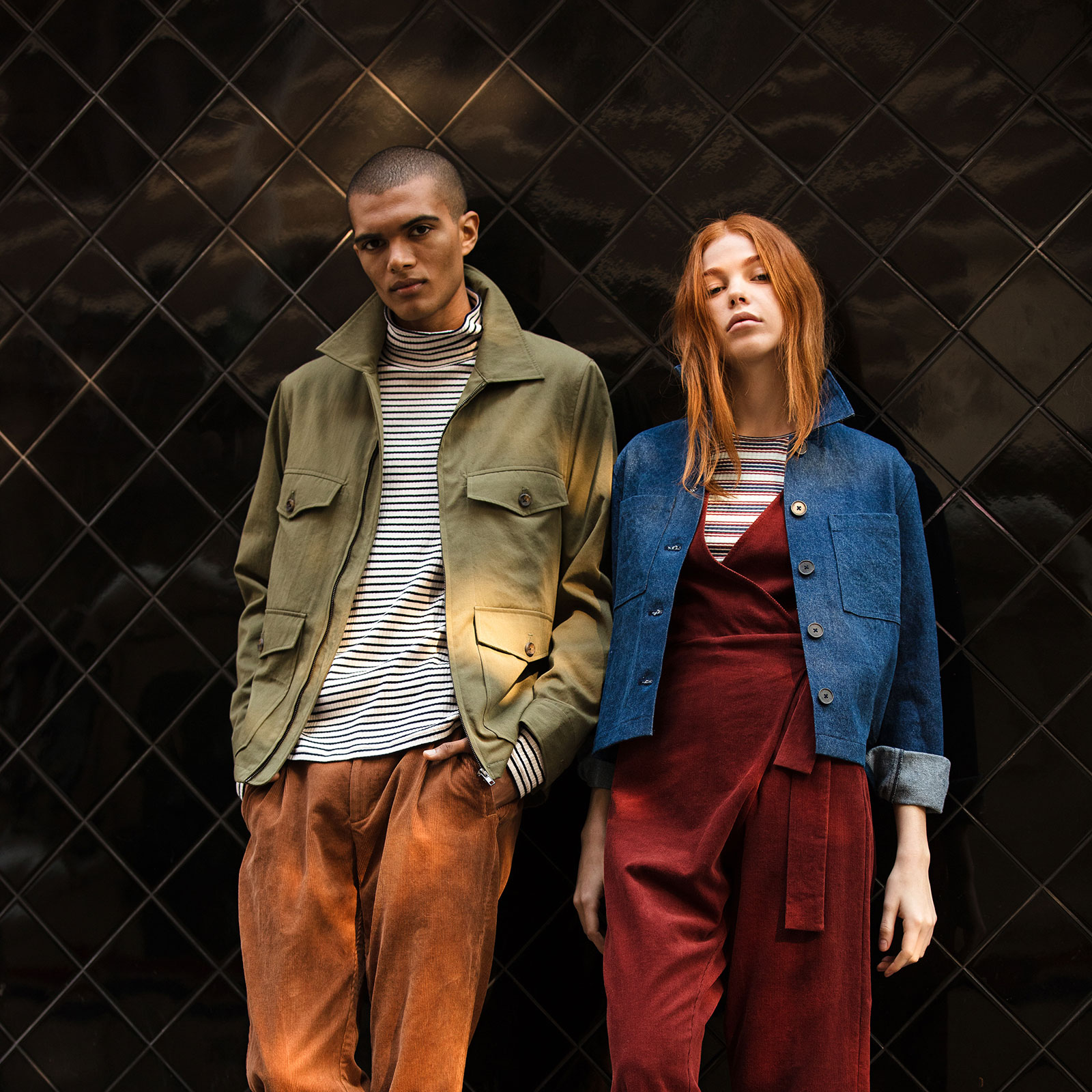 man wearing khaki jacket and orange pants leaning against a wall next to a woman wearing denim jacket and red jumpsuit