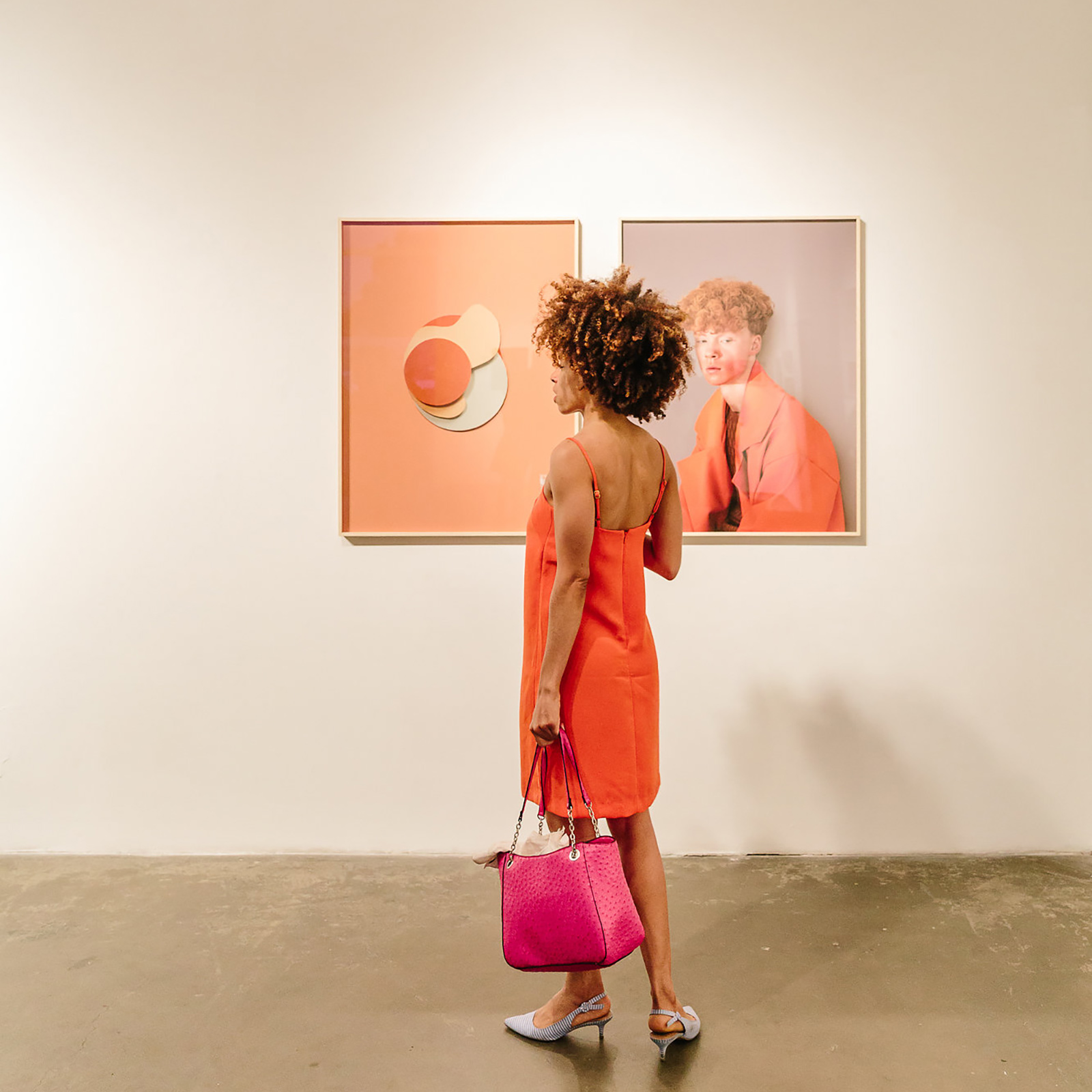 woman in orange dress holding pink bag standing in front of two paintings